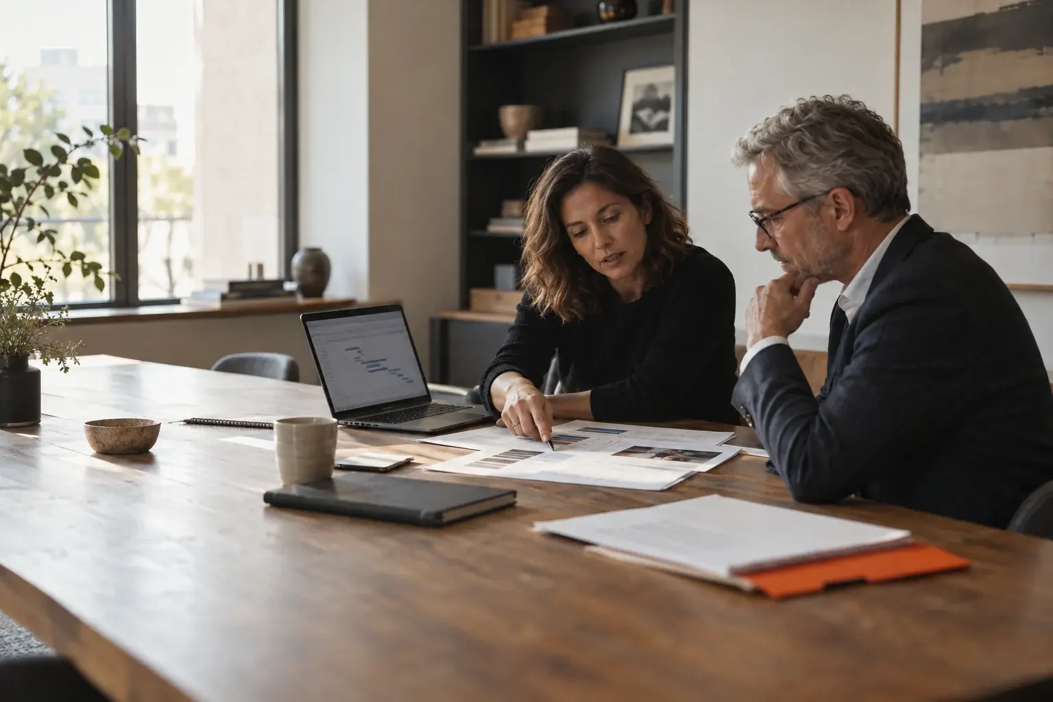 Two experienced colleagues review a proposal and delivery plan at a sunlit worktable.