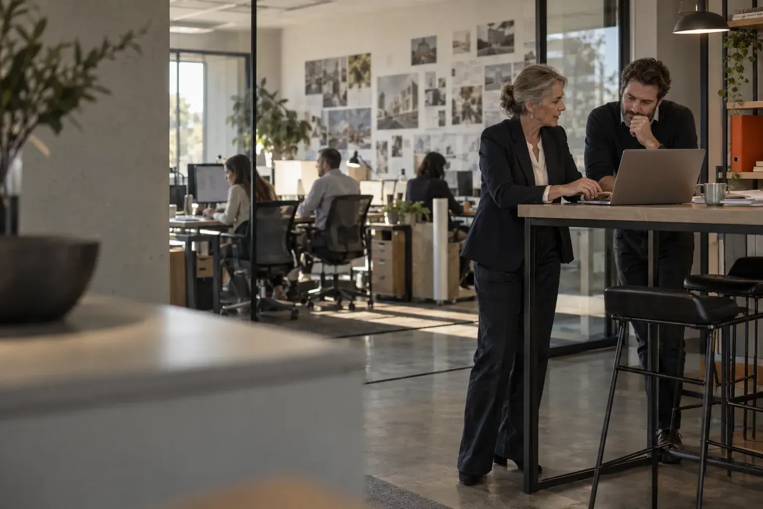 Two company leaders talk over a laptop at a high counter, with an open office behind a glass wall.